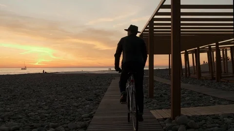 At sunset a man in a hat rides a retro bike enjoying a walk, the Sea and the 스톡 동영상 121806463