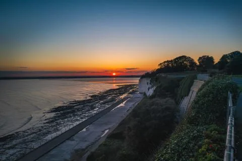 Sunset over a bay of water seen from a cliff top promenade as a person walks Stock Photos
