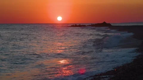 Sunset over a calm sea, with waves gently lapping at a rocky shoreline Stock Footage 318824581