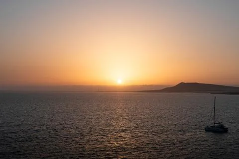 Sunset over calm waters with distant hills and a sailboat near the shore Stock Photos