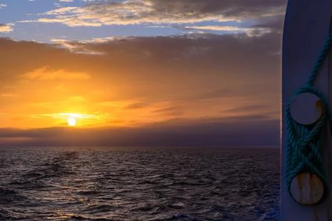 Sunset over the ocean highlighting waves and a boat's rail in quiet evening.. Stock-Fotos