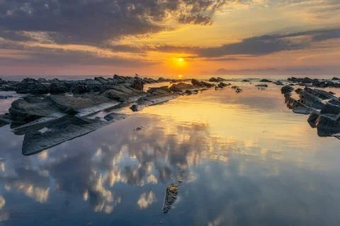 Sunset over the ocean with rocks in the foreground and calm waves. Stock Photos