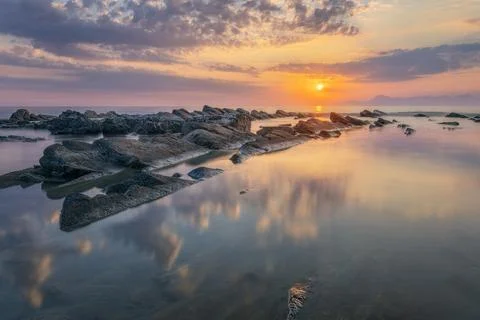 Sunset over the ocean with rocks in the foreground and calm waves. Stock Photos
