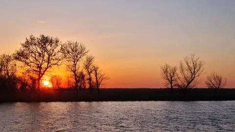 Sunset over river with silhouettes of bare trees and orange sky Stock Photos