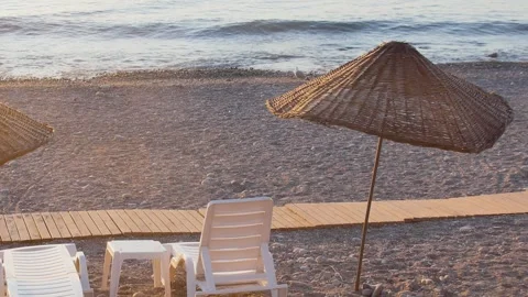 Sunset on a peaceful pebble beach featuring white sun loungers, straw umbrel Vidéo 333470462