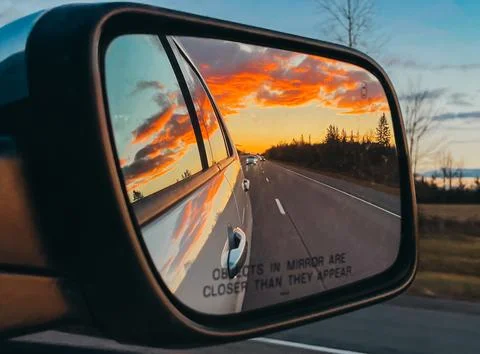 Sunset reflected in side mirror of vehicle on tree lined road. Stock Photos