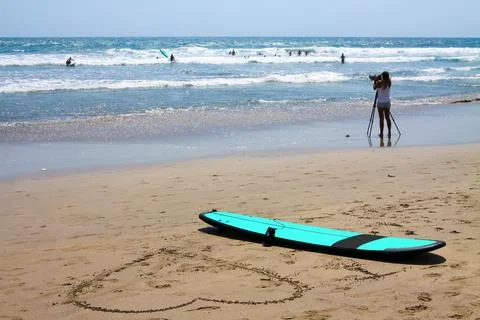 A surfboard is lying on the beach. Next to it, a large heart is drawn in th.. Stock Photos