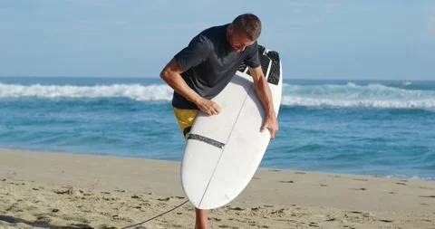 Surfer apply wax on beach with surfboard looking at ocean. Stock Footage 213613590
