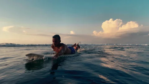 Surfer guy swims by lying on a surfboard in the ocean at sunset, clouds Stock Footage 157991513