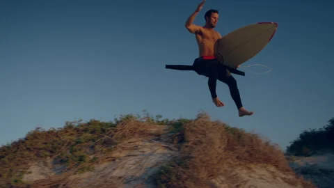 Surfer jumping off of a small sand hill at the beach Stock Footage 279783965