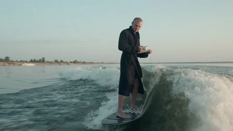 A surfer jumping on a wakeboard in a terry dressing gown with a cup Stock Footage 160371413