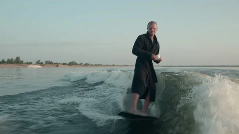 A surfer jumping on a wakeboard in a terry dressing gown with a cup Video stock 160393235