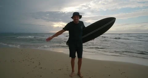 Surfer man standing on sandy beach with surfboard near ocean during strong wind. Stock Footage 276589173