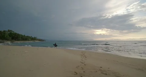 Surfer man walks on sandy beach with surfboard near ocean at rainy evening. Stock Footage 276764365