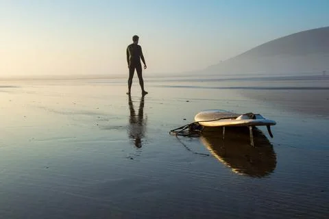 A surfer next to a surfboard on a beach reflected by the water on the sand. Stock Photos