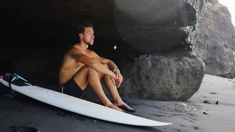 Surfer sits at cave near rocks at Bali with surfboard Stock Footage 87976541