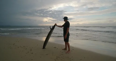 Surfer with surfboard stands on ocean beach observing waves at sunset. Stock Footage 276629461
