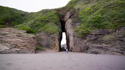 A surfer walking across the black sand towards a cave, Iglesia de Piedra, Chile. Stock Footage 345700021