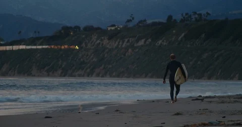 Surfer walking on beach at sunset as freight train passes by on hills above 库存影片 128669090