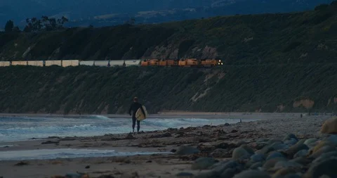 Surfer walking on beach at sunset as freight train passes by on hills above 库存影片 128709294