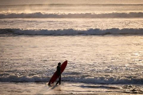 Surfer walking through shallow waves carrying red surfboard Foto stock