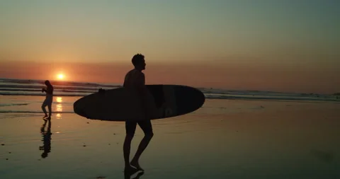Surfer walks along beach at sunset 스톡 동영상 138328989