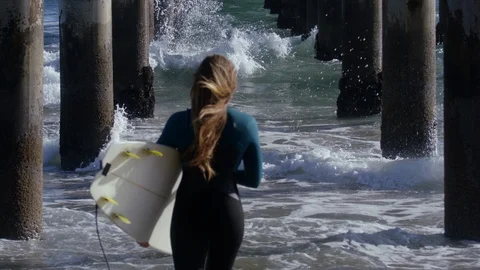 Surfer Walks Out Into Waves, Under Pier, Her Hair Blows In Breeze (Slow Motion) Stock Footage 92493624