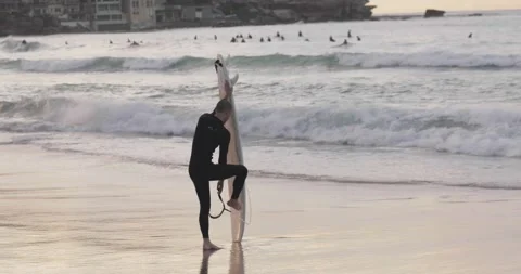 Surfer wraps leg strap around board as he exits the surf during sunrise Stock Footage 137624753