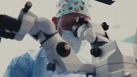 Surgeon in blue scrubs and patterned cap focuses on surgical microscope during Stock Footage 337286112