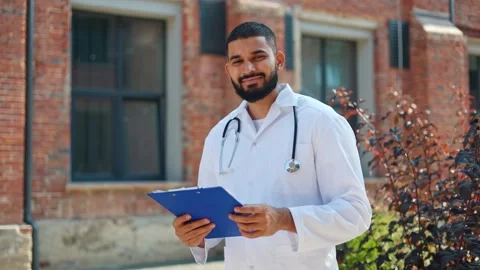 Surgeon of indian ethnicity checking medical records and smiling at camera Stock Footage 250076341