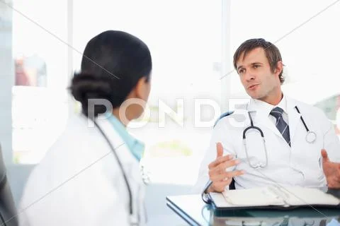 Surgeon Sitting At Desk With A Notebook While Talking To A Colleague