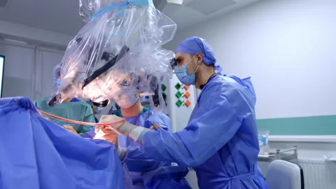 Surgeons in blue uniforms sit at microscope performing operation. Stock Footage 292147557