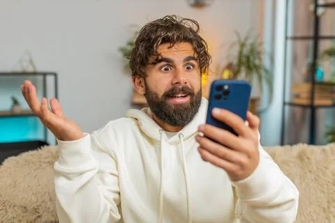 Surprised Indian bearded man using cellphone winning lottery sitting on sofa Stock Photos