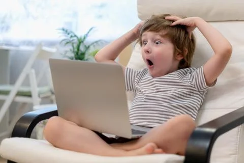 Surprised kid boy sit on computer using laptop and having a video call. Child Stock Photos