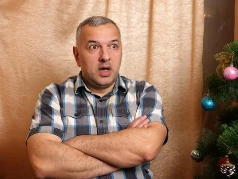 Surprised man with crossed arms sitting beside a Christmas tree, reacting emo Stock Photos