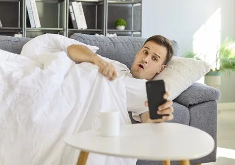 Surprised man on sofa under blanket checks phone alarm after waking late Stock Photos