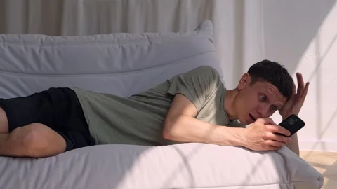 Surprised man staring at phone screen with strong emotion while relaxing at home Stock Footage 311445854