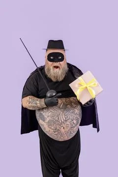 Surprised mature man with overweight in Zorro suit points onto gift box on Stock Photos