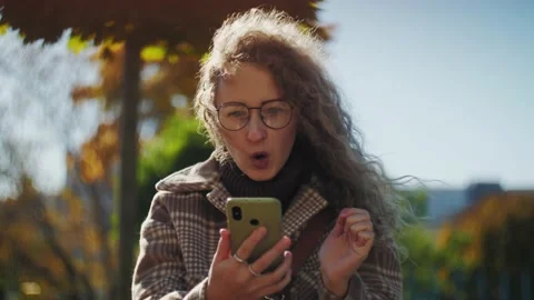 Surprised Woman using phone sitting in the park.Curly girl with glasses holding Stock Footage 220766434