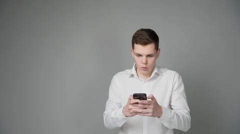 A surprised young guy looks at the screen of his smartphone. On a gray backgr Stock Photos