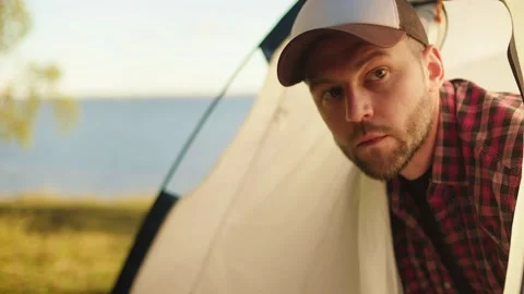 A surprised young tourist in a cap gets out of a tent looks around and leaves it Stock Footage 148189581