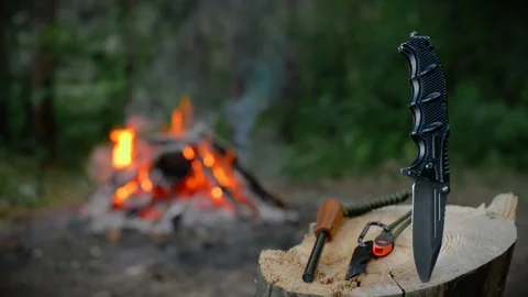 Survival knife and fire striker flint on wood log with campfire in forest Stock Footage 341878498