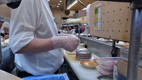 Sushi Chef Preparing Plates on Conveyor Belt at Busy Kaiten Sushi Restaurant in Stock Footage 337745649