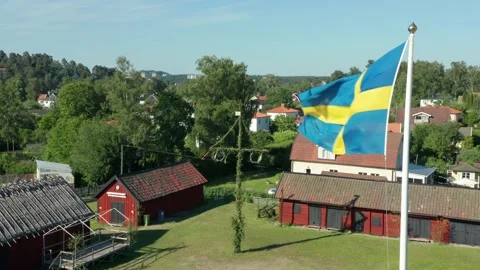 Swedish flag and midsummer may pole in background. Small red farm on countryside Stock Footage 198652361