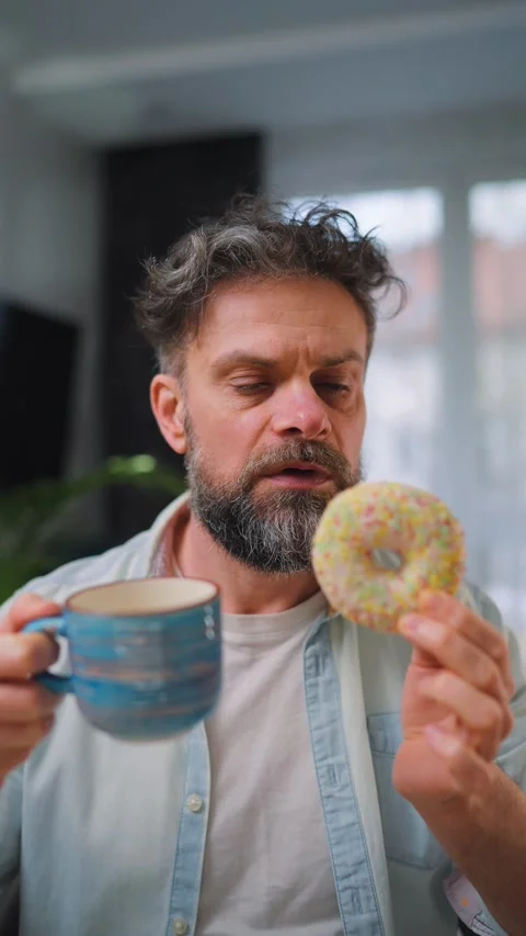 Sweet addiction concept. A man eats a donut and drinks coffee. Vertical Video stock 342958044