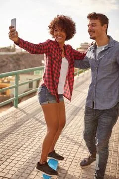 Sweet couple taking a selfie while the girl is balancing on a skateboard Stock Photos
