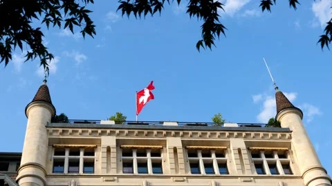 Swiss Flag Waving on an Old Building with Tower and Tree Branch Against Blue Sky Stock Footage 345786335