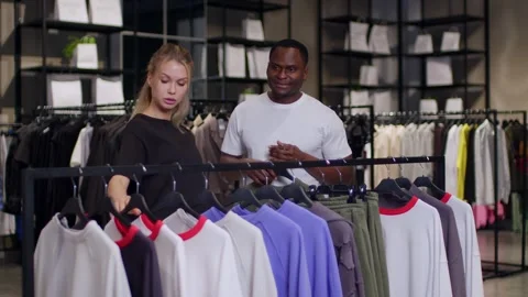 In a T-shirt store, a light-skinned consultant helps a dark-skinned man choose 库存影片 263394500