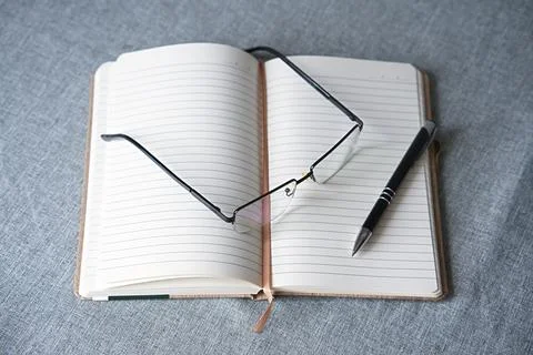 On the table are an open lined notebook, a black pen, and thin-framed glasses Stock Photos