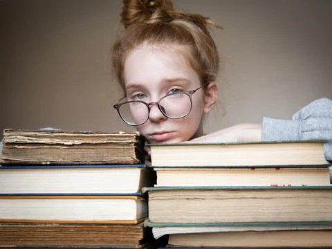 On the table are two large stacks of books on which lay a girl with red hair in Stock Photos
